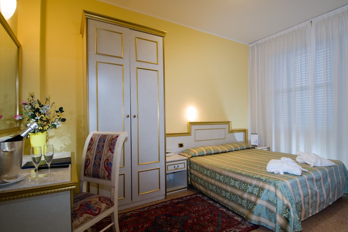 A hotel room with a bed, a nightstand, a chair, a wardrobe, a mirror, and a window with curtains.