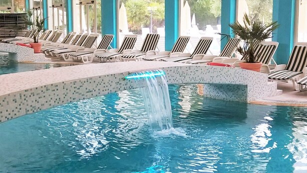 A swimming pool with a waterfall feature, surrounded by lounge chairs and potted plants. - Hotel Terme Internazionale (Abano Terme)