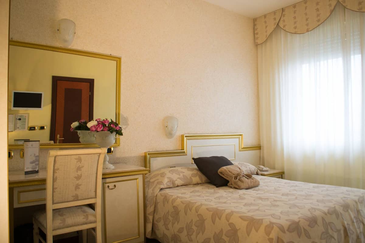 A hotel room with a bed, a desk with a chair, a mirror, and a window with curtains.