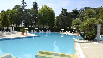 Una piscina con tumbonas y sombrillas, rodeada de árboles y un suelo de baldosas.