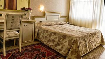 A hotel room with a bed, a desk, a chair, a nightstand, a vase with flowers, a wall-mounted light, and a red patterned rug.