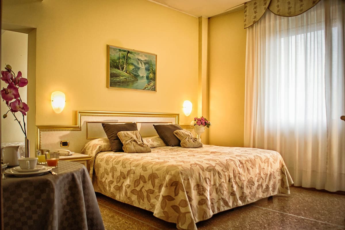 A hotel room with a bed, a painting on the wall, a nightstand, a table with a vase of flowers, and a window with curtains.