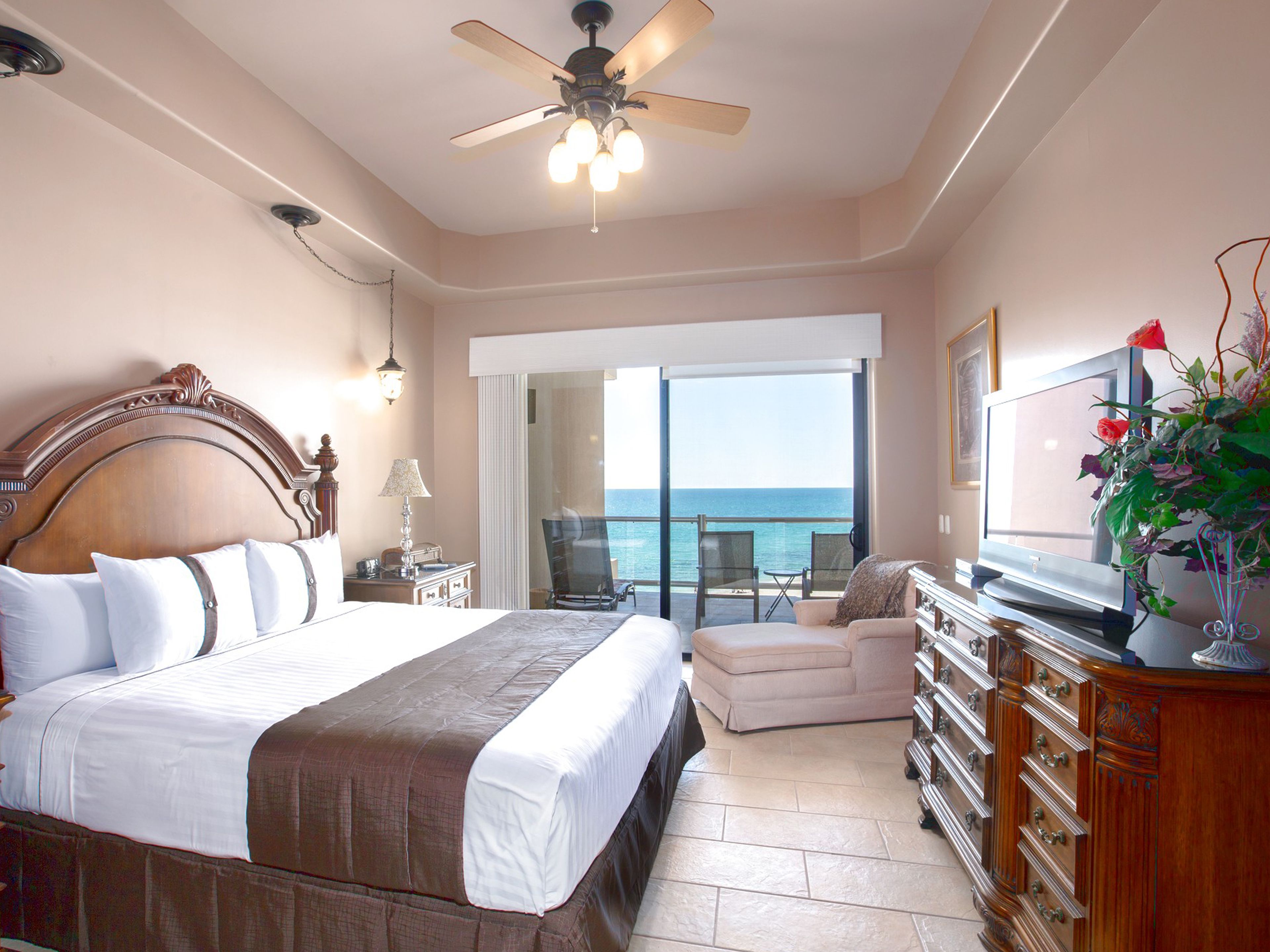 A bedroom with a large bed, a ceiling fan, a TV, and a view of the ocean.