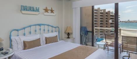 Habitación de hotel con cama, mesita de noche, lámpara, silla y vista a la playa y al océano.