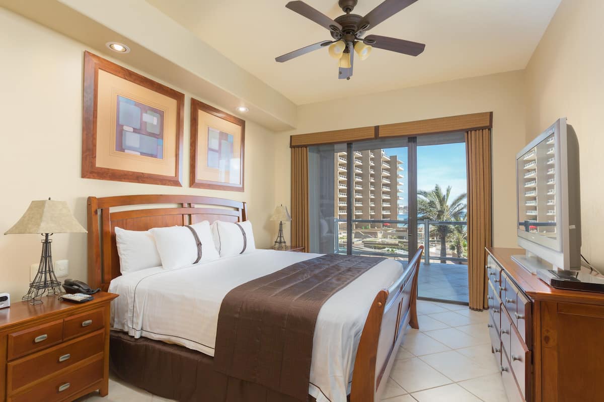 Habitación de hotel con cama, escritorio, silla, televisor, ventilador de techo y balcón con vistas a edificios y palmeras.
