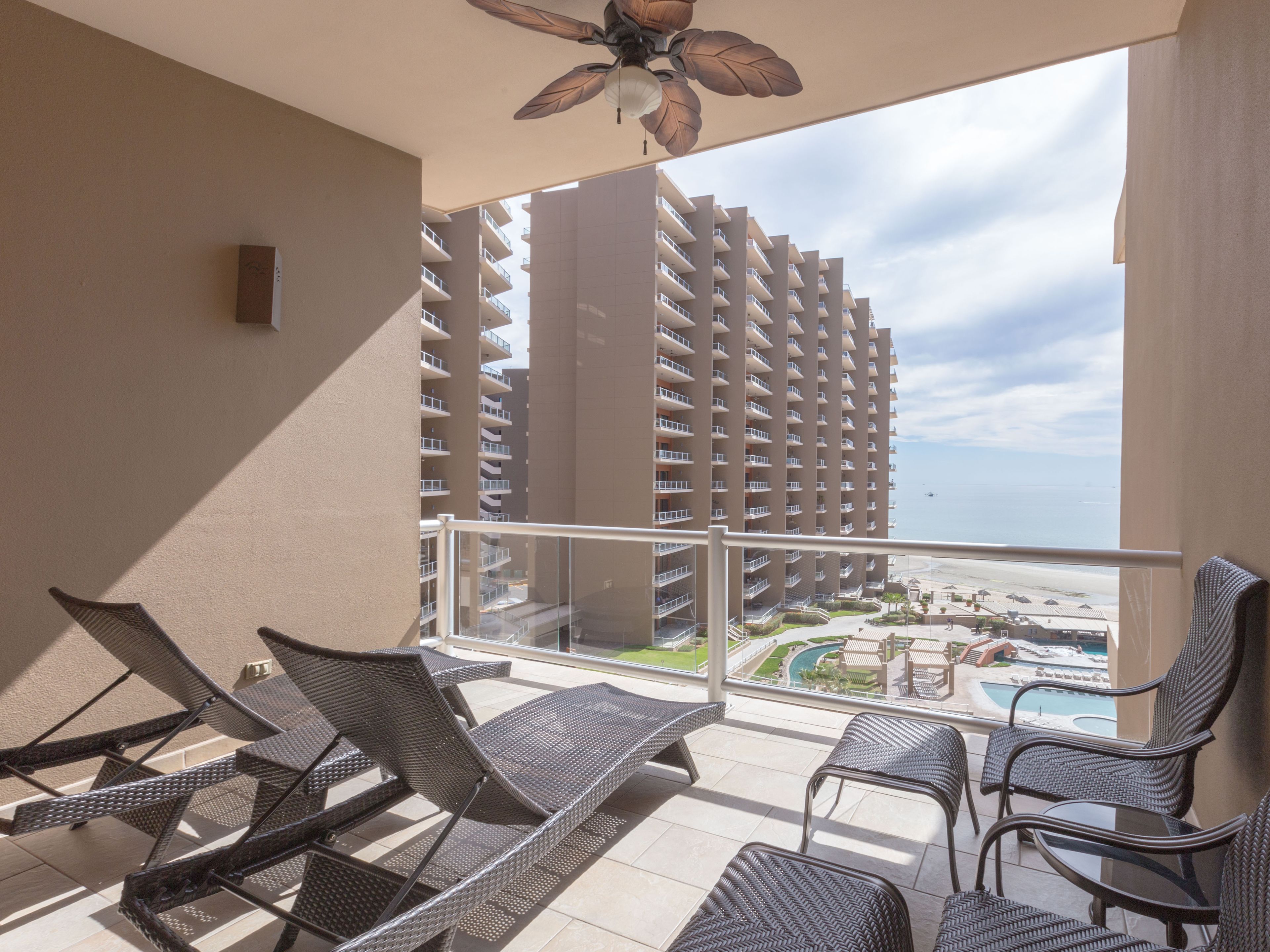 A balcony with wicker chairs, a ceiling fan, and a view of a high-rise building and beach.