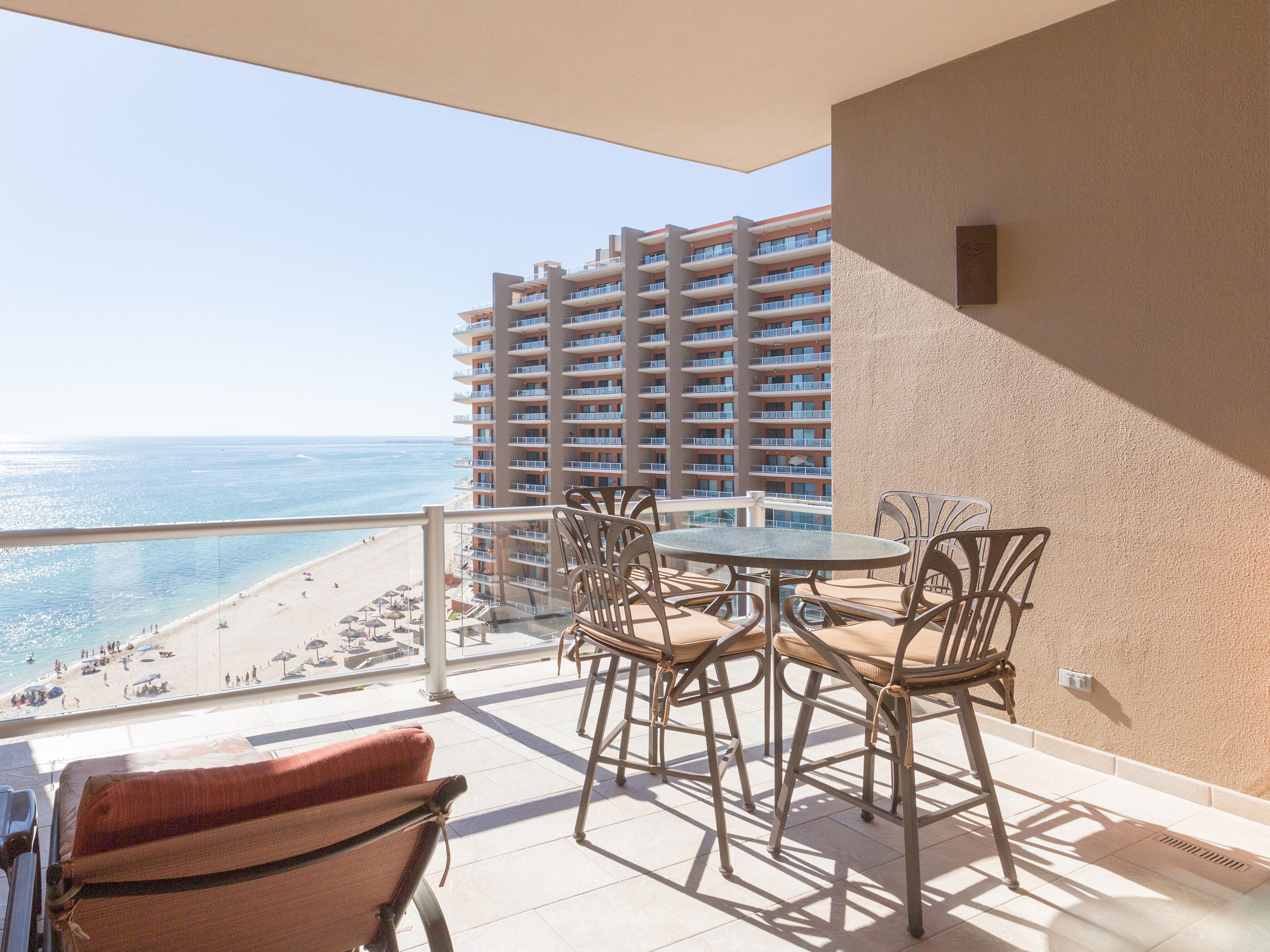A balcony with a view of the beach and a tall building. There is a table with chairs and a sofa.