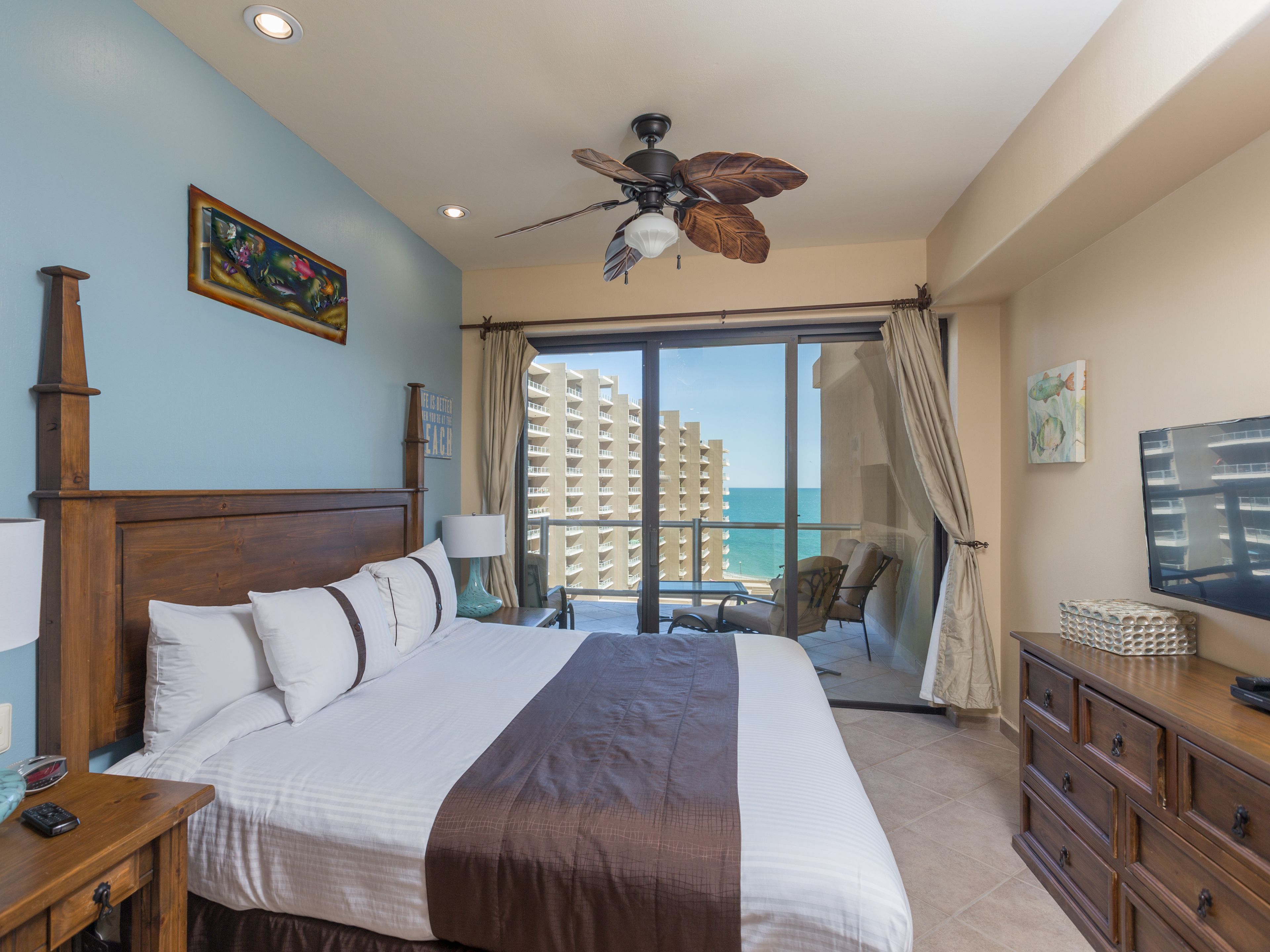 Habitación de hotel con una cama grande, ventilador de techo, televisor, escritorio y vistas al océano y edificios.