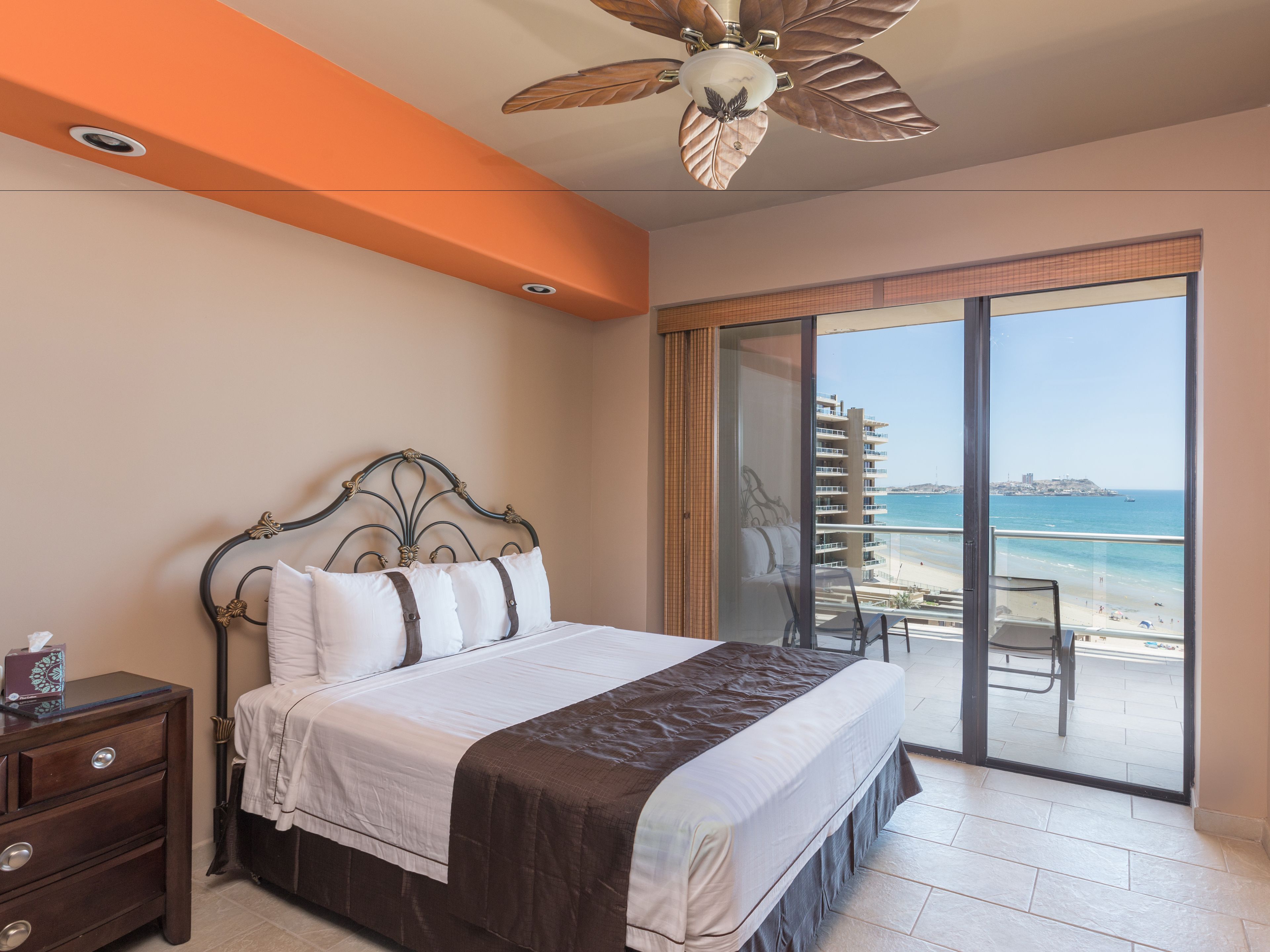A bedroom with a bed, a nightstand, a ceiling fan, and a balcony with a view of the beach and ocean.
