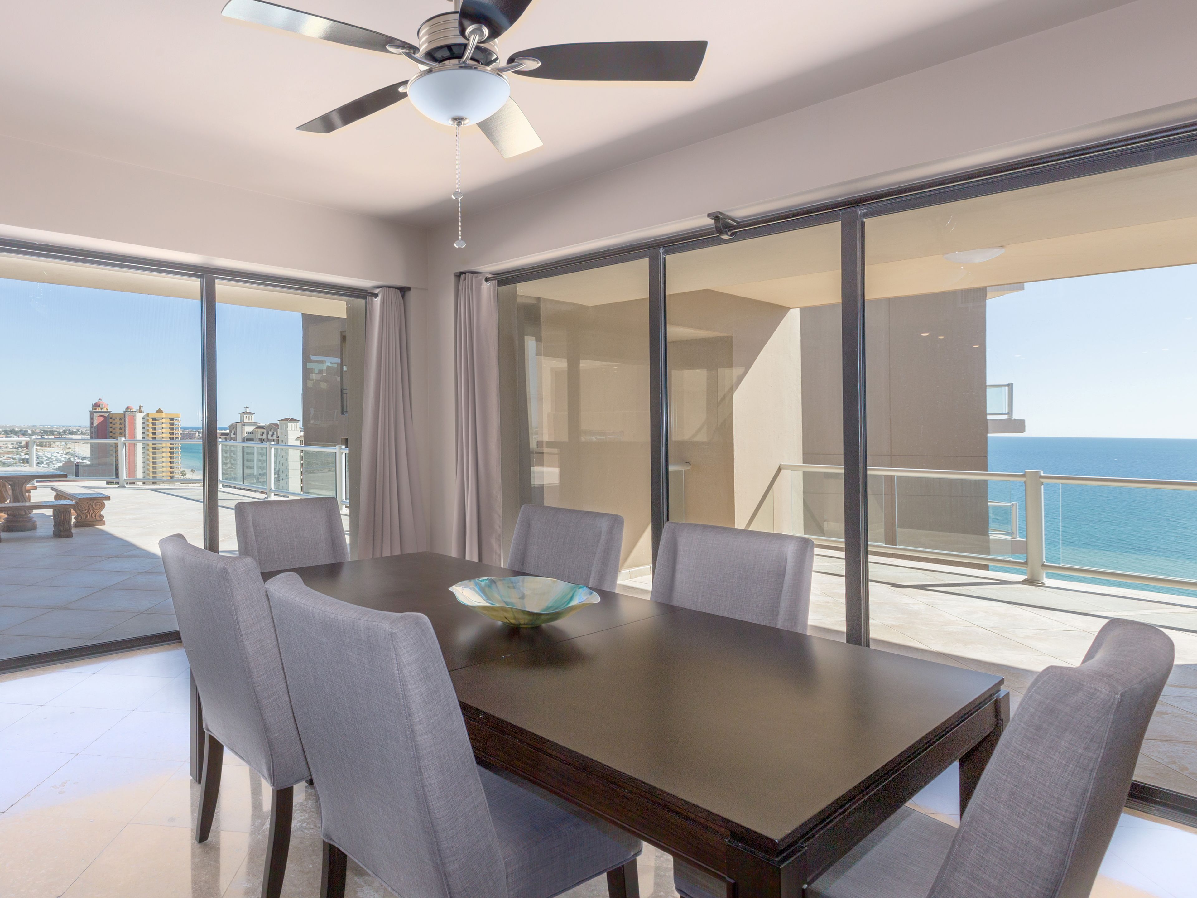 Un balcon avec une table et des chaises pour prendre les repas, un ventilateur de plafond et une vue sur l’océan et les bâtiments.