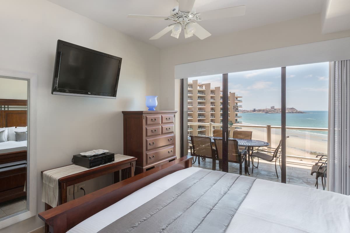 Habitación de hotel con cama, escritorio, televisor, cómoda y balcón con vista a la playa y al océano.