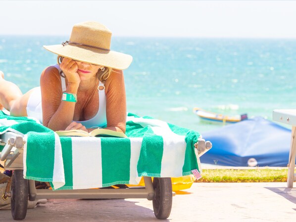 A person reclining on a beach chair with a green and white striped towel, reading a book, with a clear view of the ocean and boats in the background.