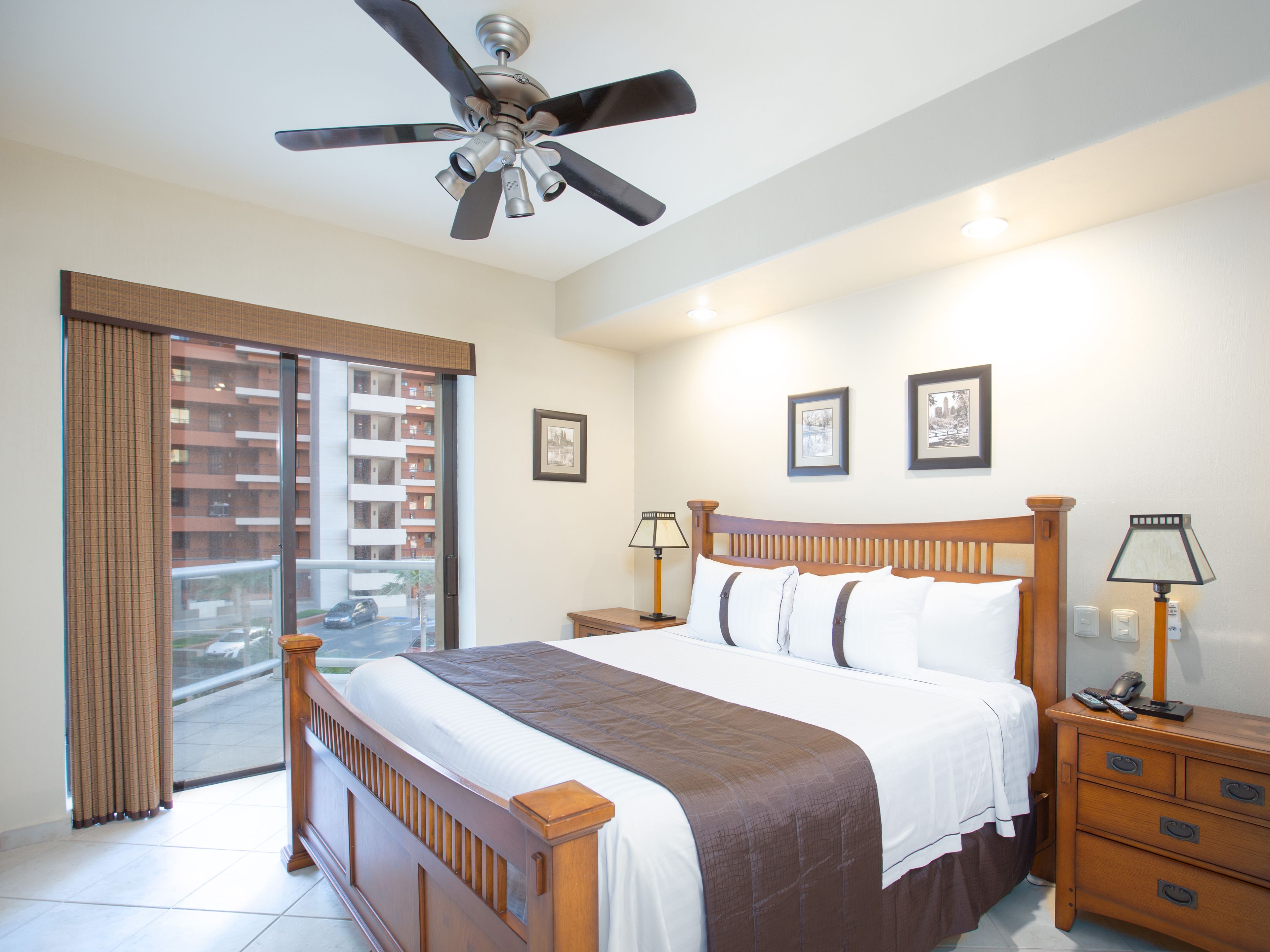 A hotel room with a bed, bedside tables, a ceiling fan, and a view of a building through the window.