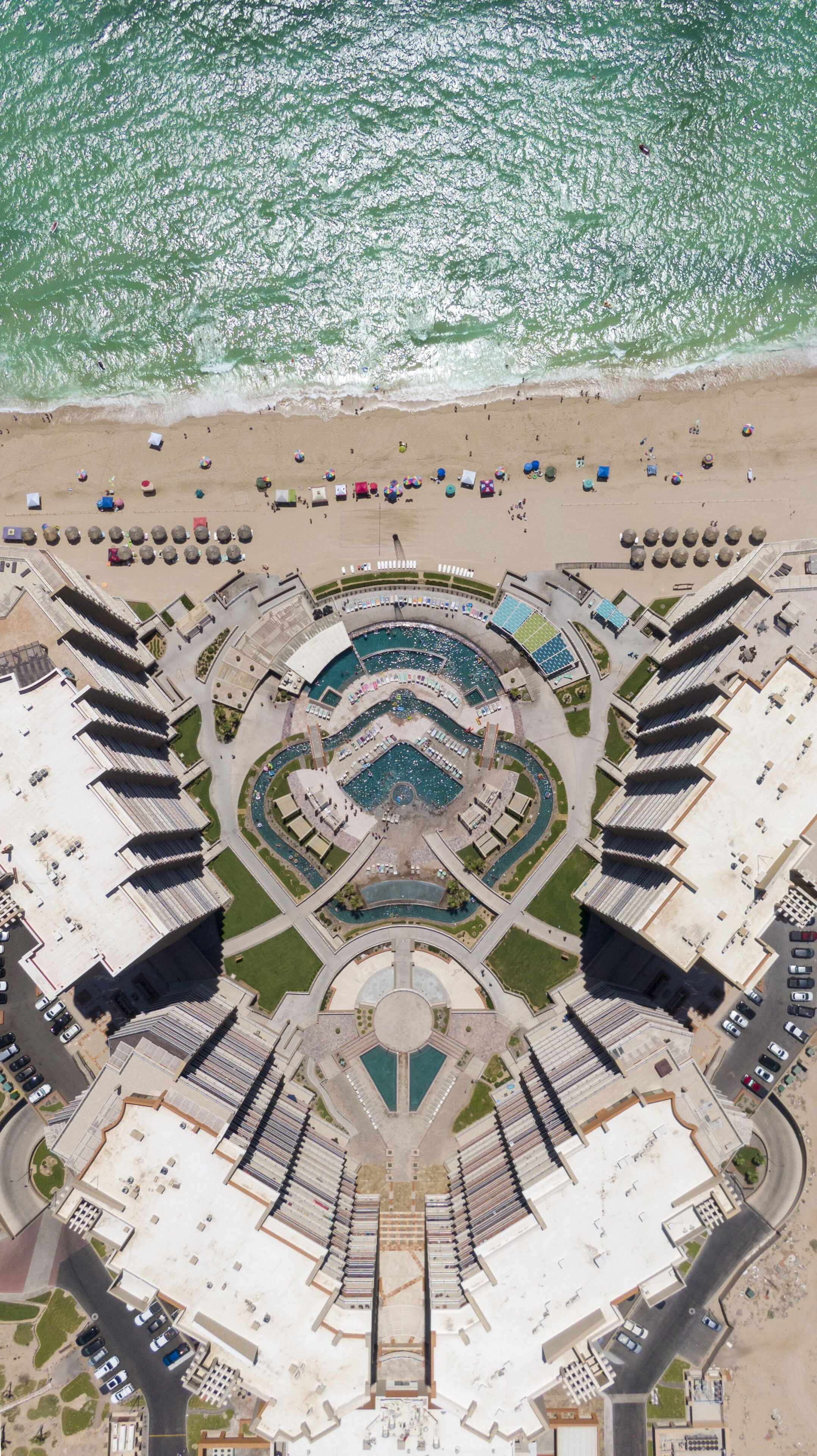 Luftaufnahme eines Küstenresorts mit zentralem Pool, Strandstühlen und einem Parkplatz.