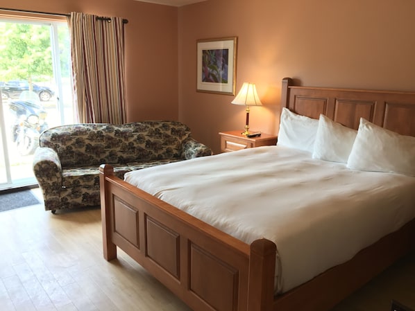 A hotel room with a wooden bed, a floral-patterned armchair, a nightstand with a lamp, and a door leading to a parking area.