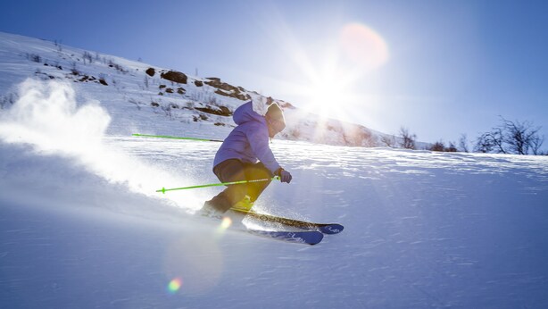 A person skiing on a snow-covered slope with a clear sky and sun in the background.