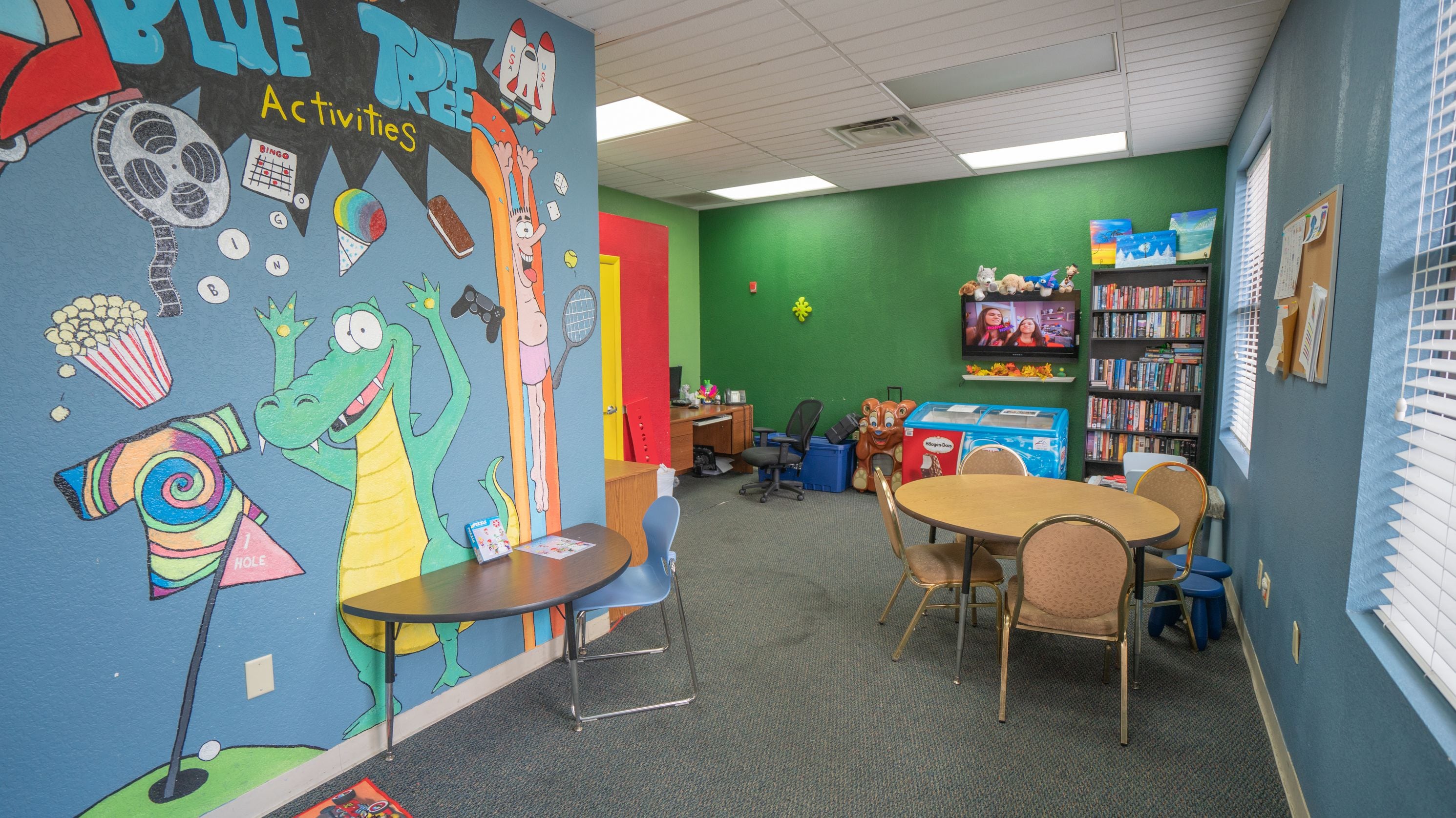 Una sala d'activitats infantil amb un mural colorit, una taula amb cadires, una prestatgeria i una màquina expenedora.