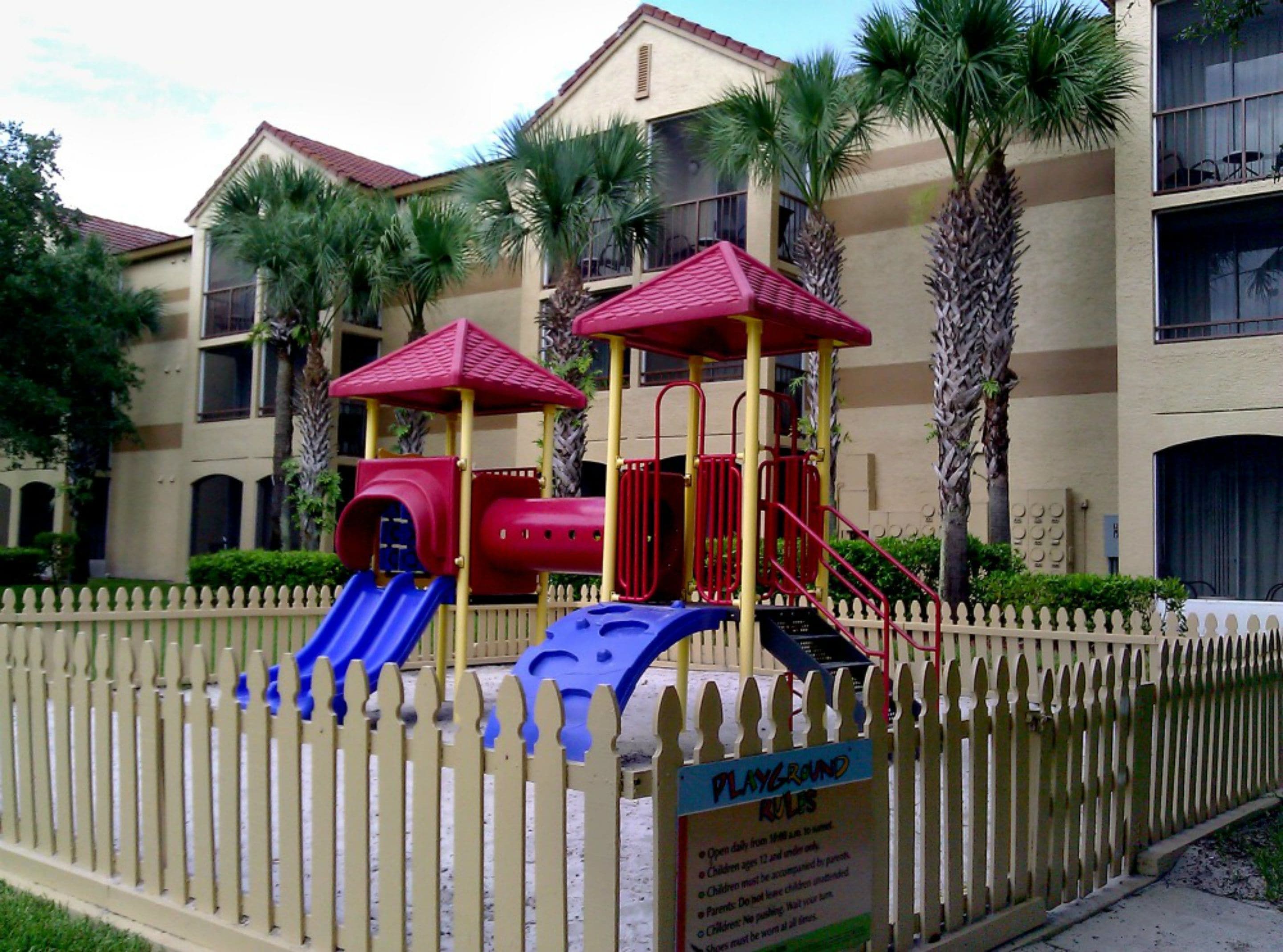 A playground with a slide and climbing structures, surrounded by a white picket fence, in front of a building with balconies and palm trees.
