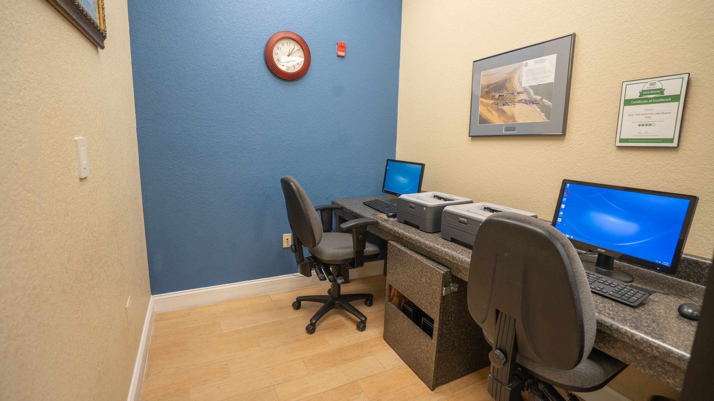 Un bureau d’ordinateur avec deux écrans, un clavier et une imprimante. Il y a une chaise devant le bureau et une horloge au mur.