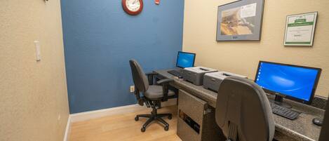 A computer desk with two monitors, a keyboard, and a printer. There is a chair in front of the desk, and a clock on the wall.