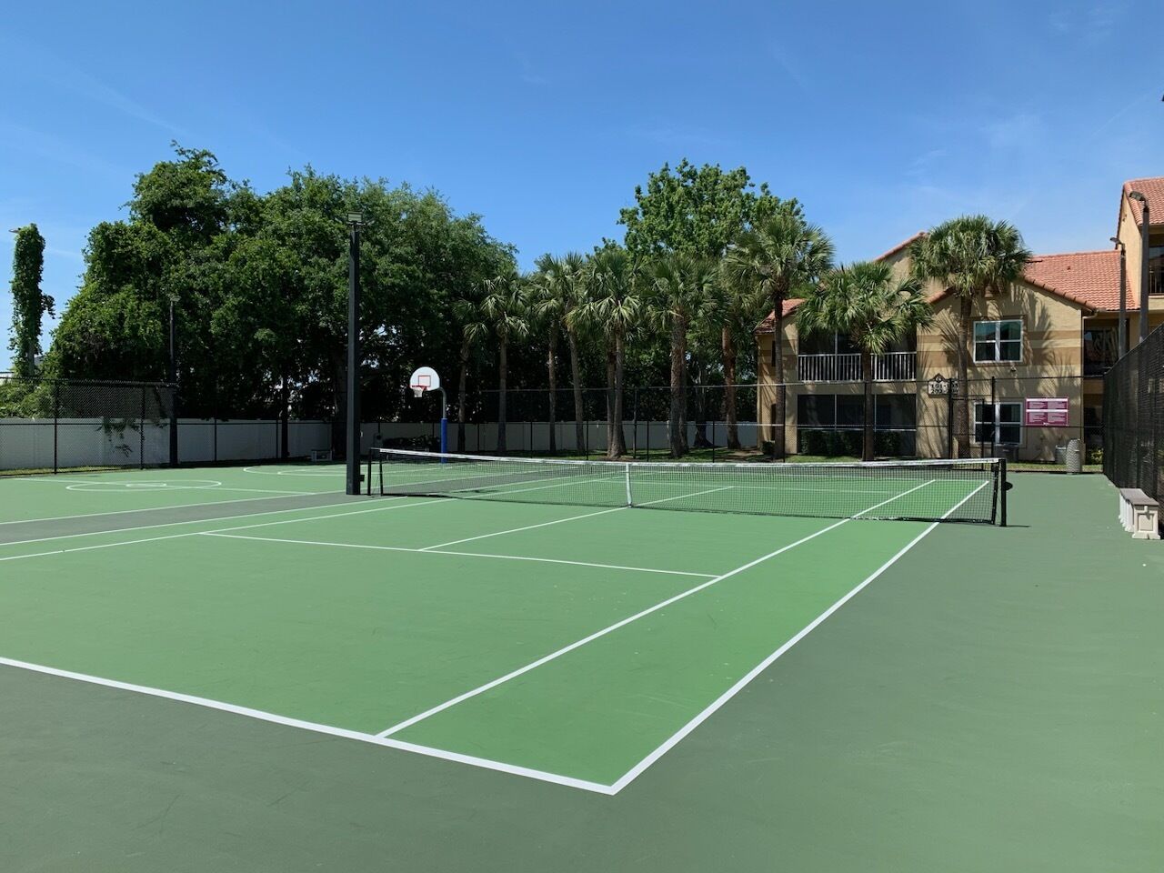 Ein Tennisplatz im Freien mit Netz, umgeben von Bäumen und einem Gebäude im Hintergrund.
