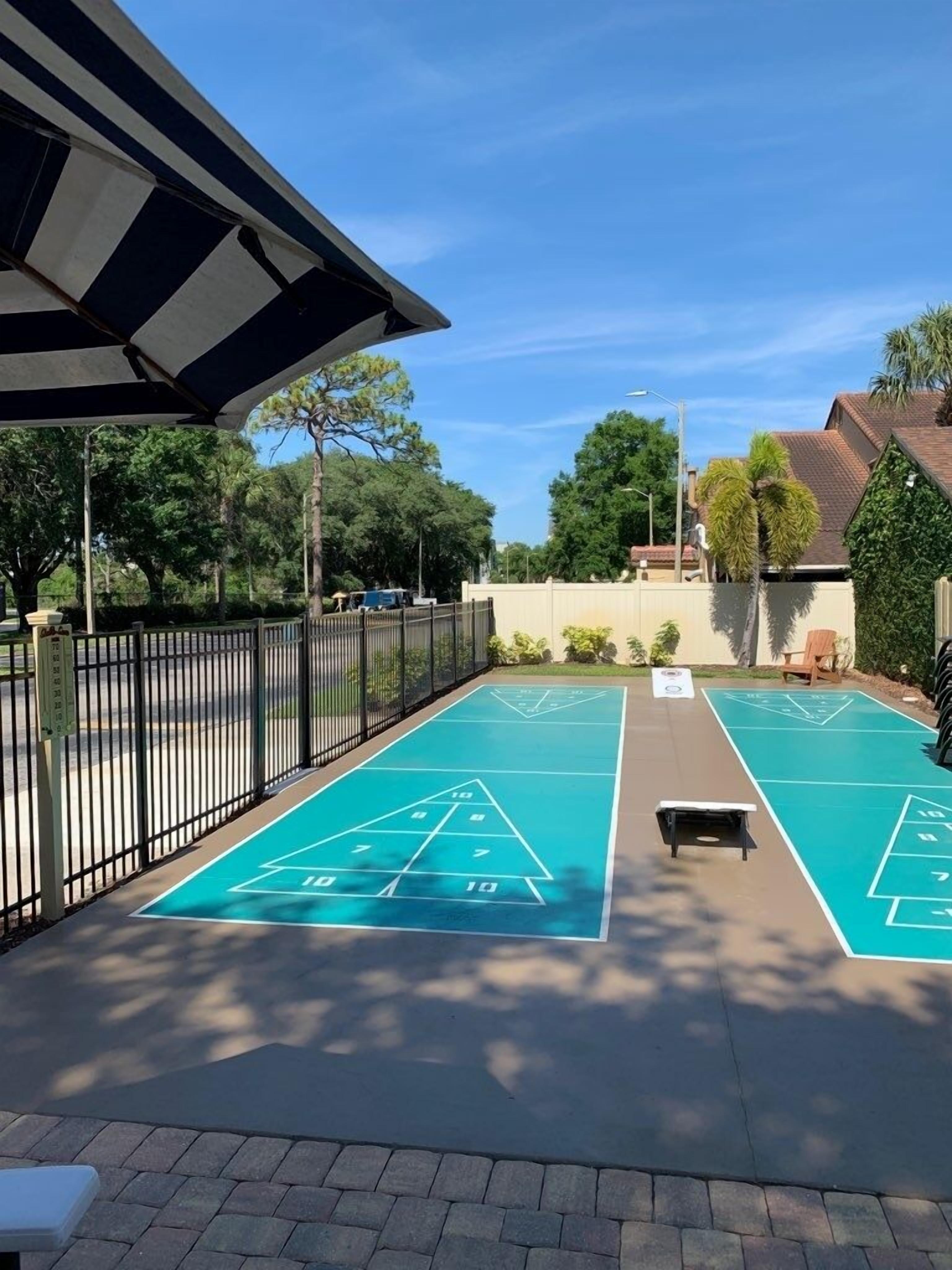 A shuffleboard court with a metal fence, a bench, and trees in the background.