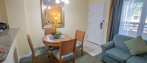 Comedor con una mesa redonda de madera, dos sillas y un jarrón con flores amarillas. Se ve un sofá azul en primer plano, y una puerta al fondo.