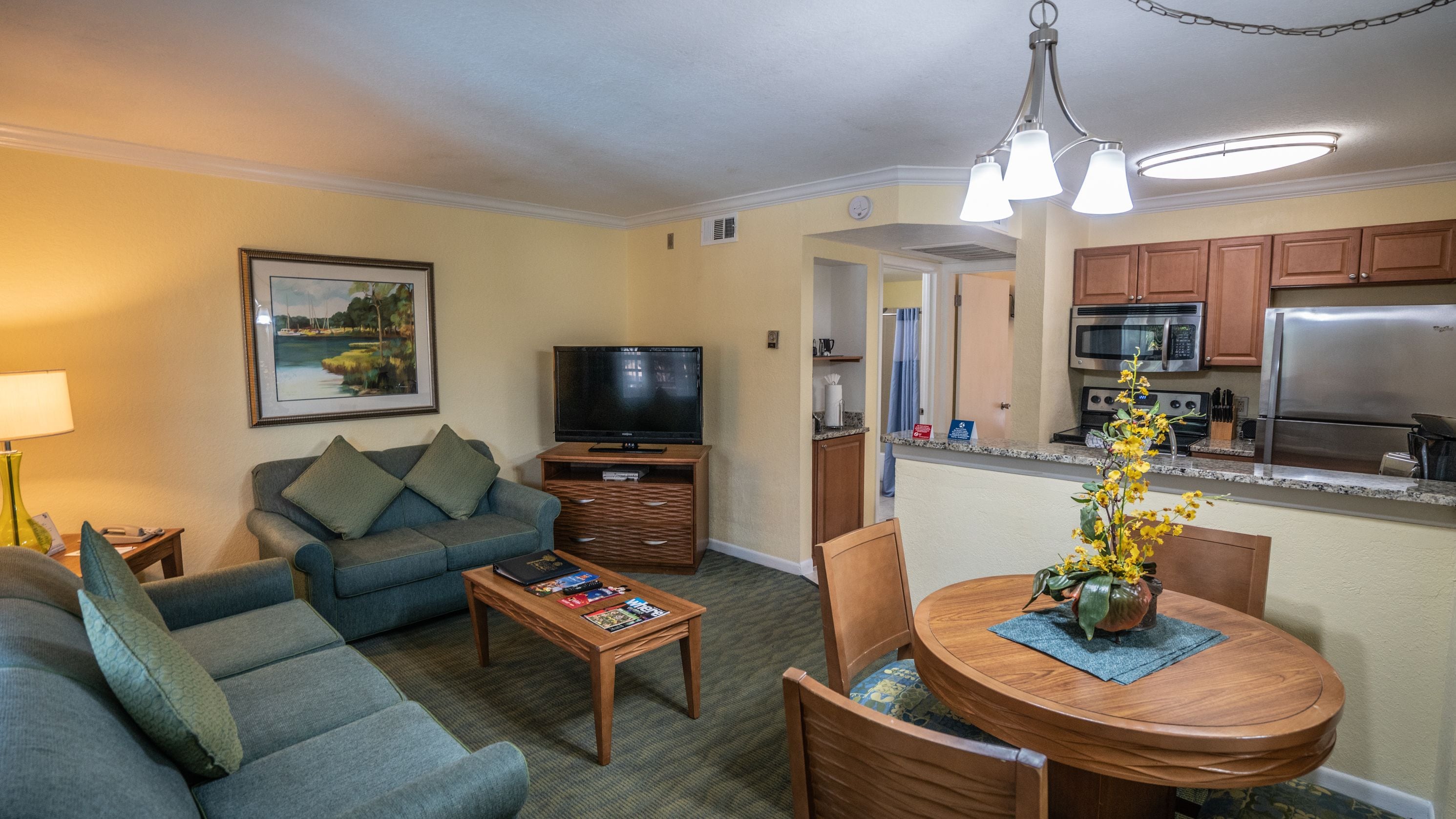 A living room with a sofa, coffee table, and TV. There's a dining table with chairs and a flower vase. The kitchen is visible with cabinets and appliances.