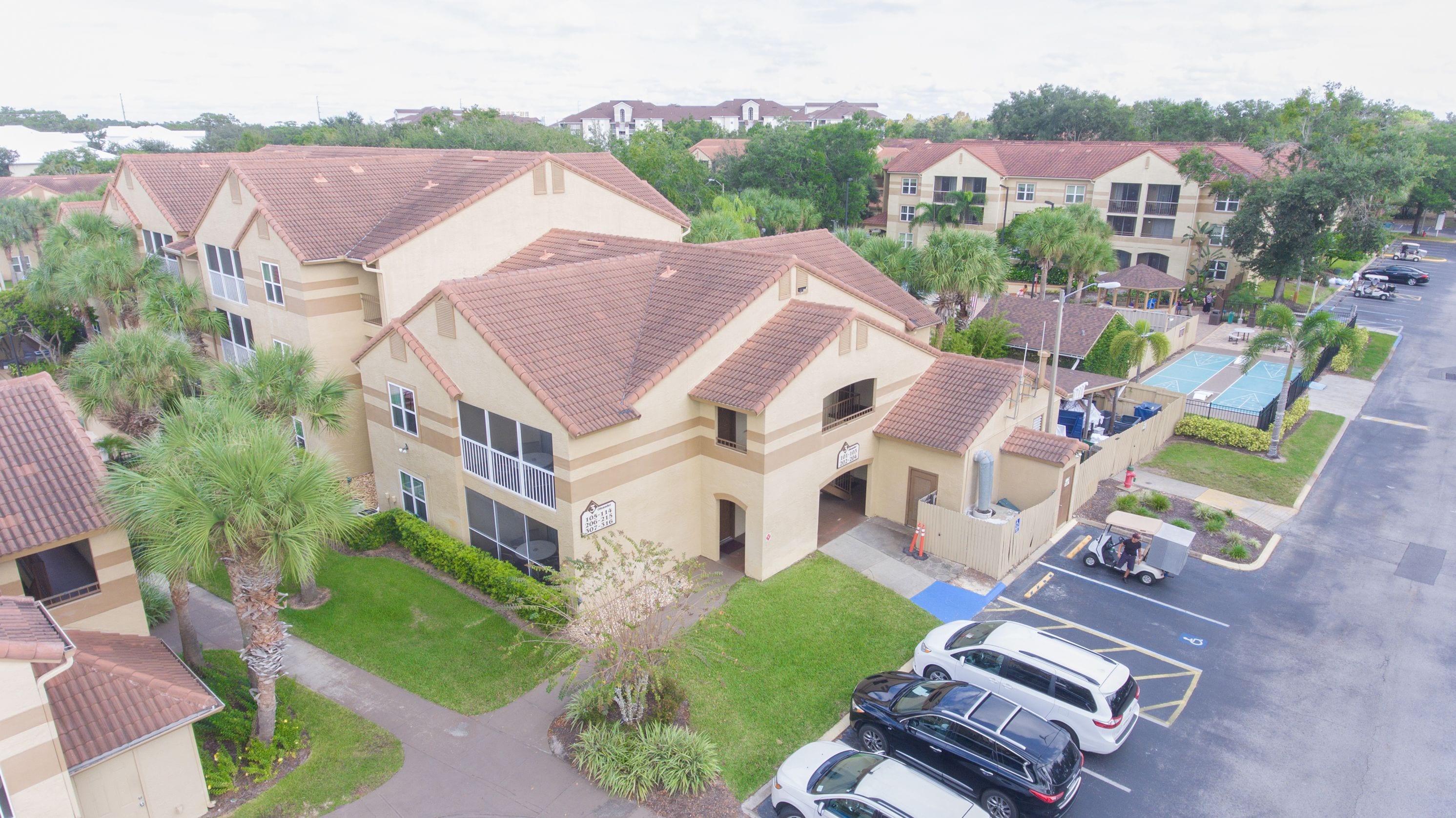 A residential building with a tiled roof, multiple balconies, and a parking area with cars.