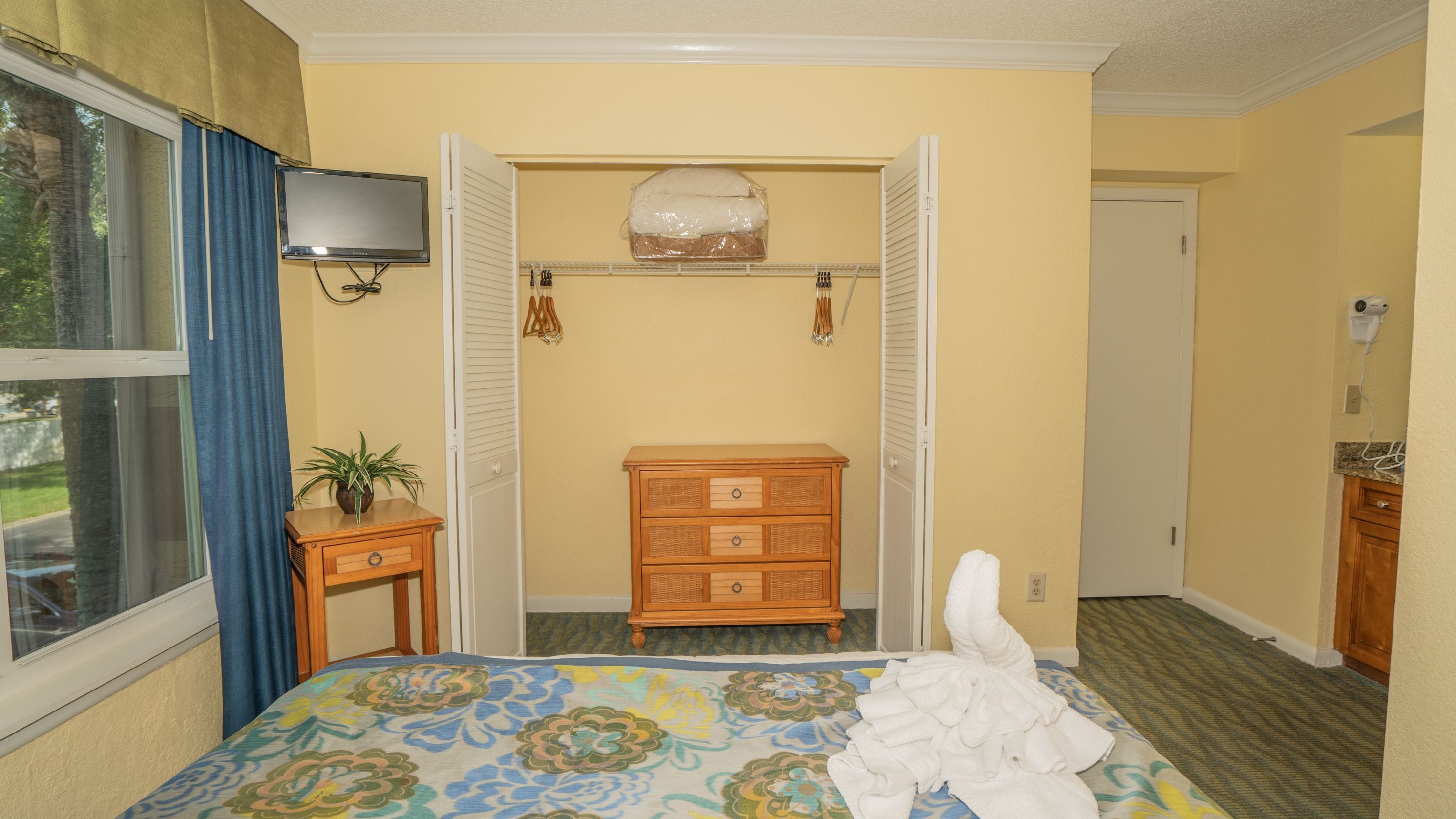 Una habitación de hotel con cama, cómoda de madera, televisor y ventana con cortinas.