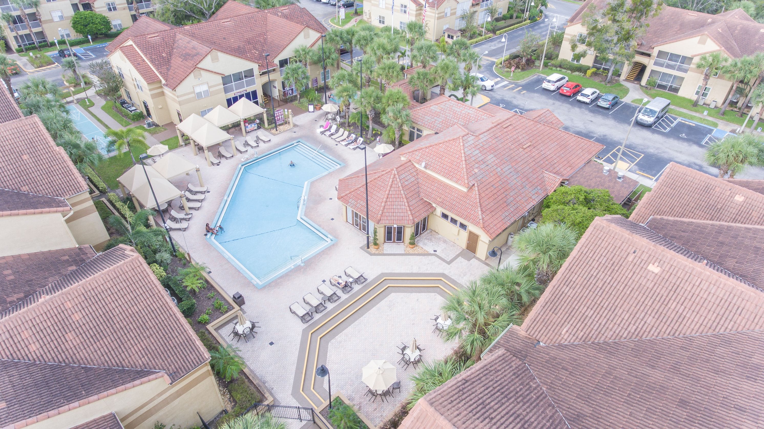 A residential area with a swimming pool surrounded by lounge chairs and umbrellas, a parking lot, and houses with red-tiled roofs.