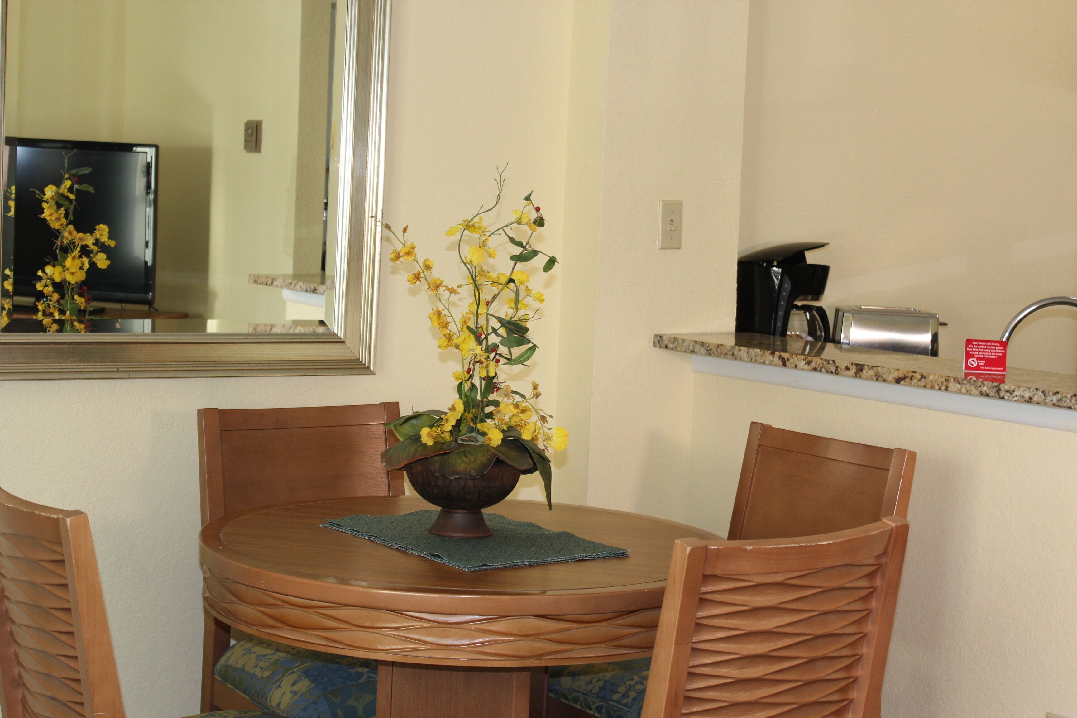 A dining area with a round wooden table, two chairs, a vase with yellow flowers, a mirror, a television, a coffee maker, and a countertop with a toaster.