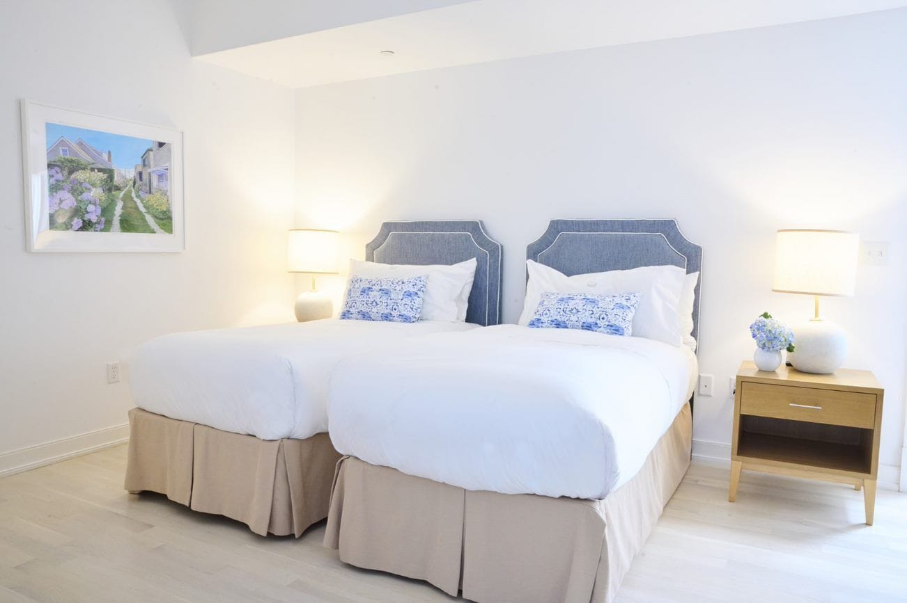 Un lit double avec une tête de lit grise, deux tables de chevet avec des lampes et un tableau encadré au mur.