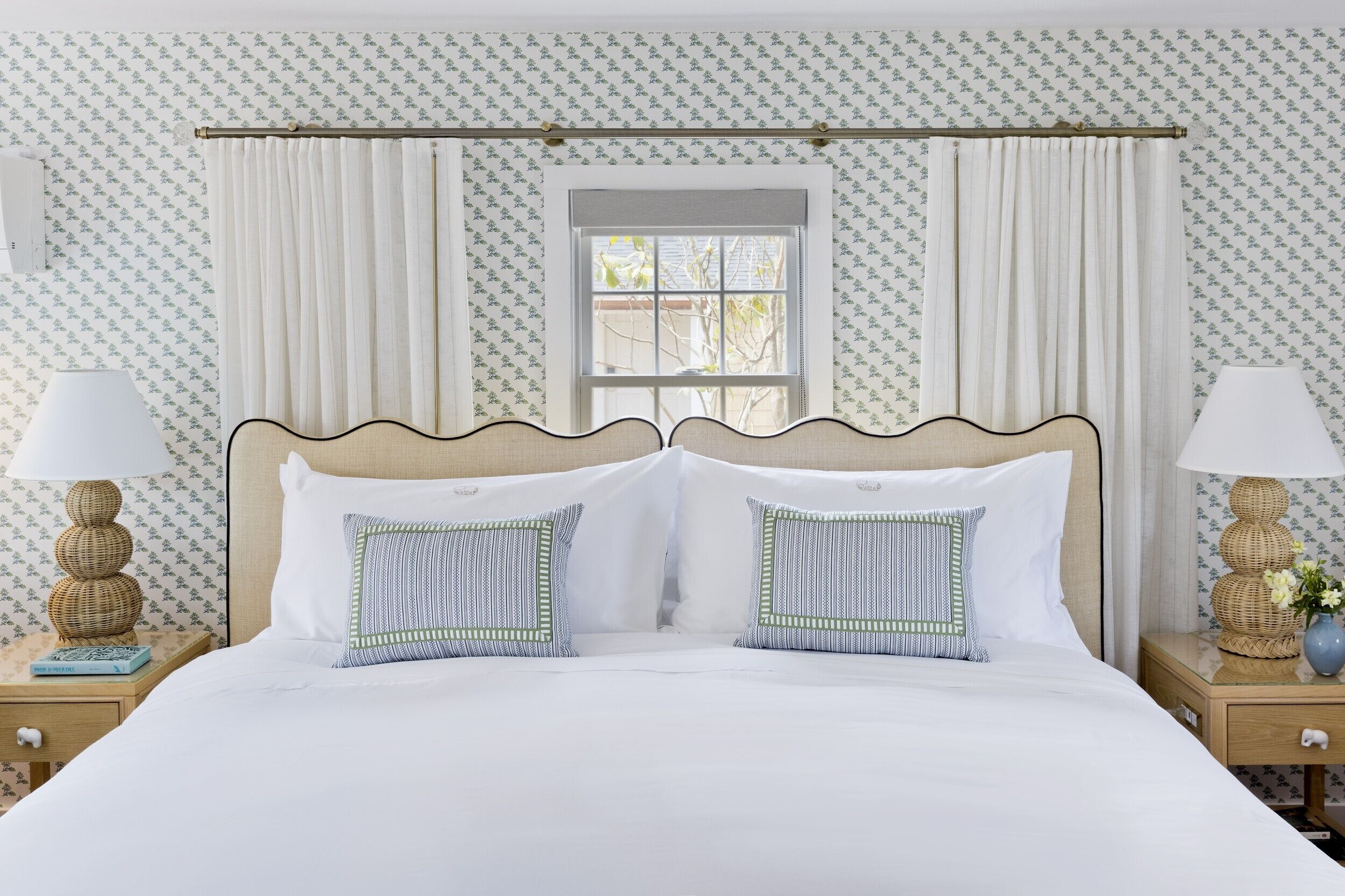Una cama bien hecha con sábanas blancas y dos almohadones decorativos, flanqueada por dos mesitas de noche con lámparas y un pequeño jarrón con flores.
