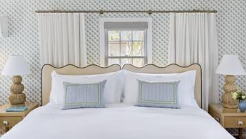 Una cama bien hecha con sábanas blancas y dos almohadones decorativos, flanqueada por dos mesitas de noche con lámparas y un pequeño jarrón con flores.