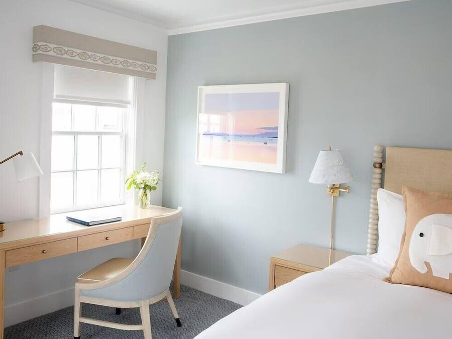 A hotel room with a desk, chair, bed, bedside table, lamp, and a framed picture on the wall.