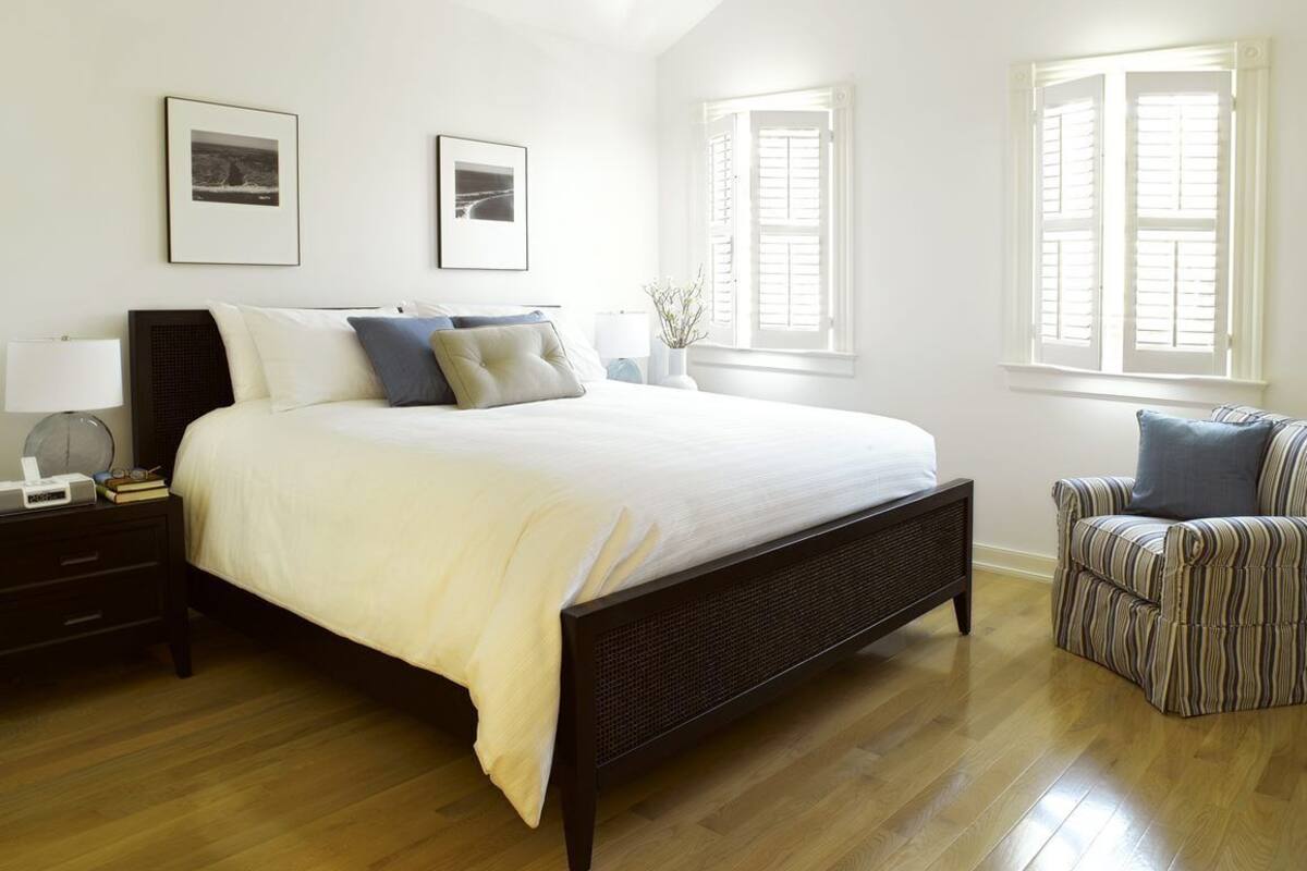 A bedroom with a large bed, a nightstand, a chair, and two windows with shutters.