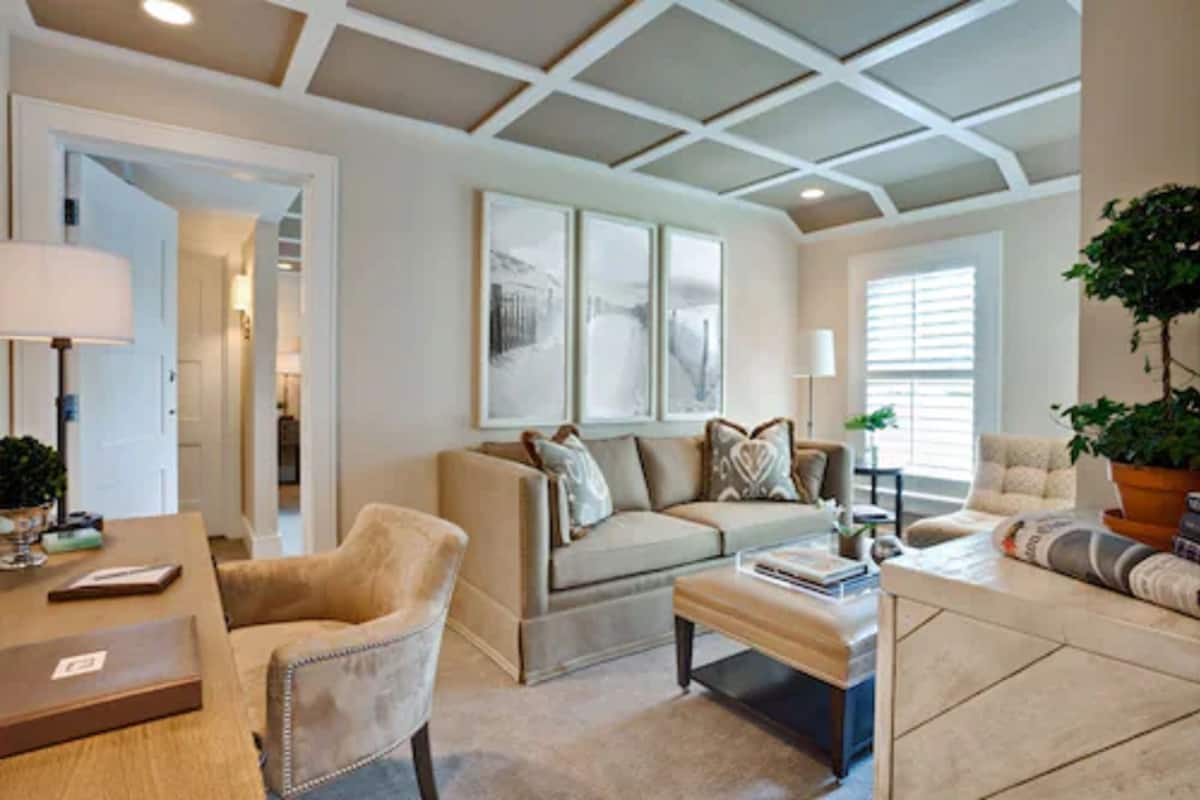A living room with a sofa, armchairs, a coffee table, and a TV. There are framed pictures on the wall and a plant on the right.