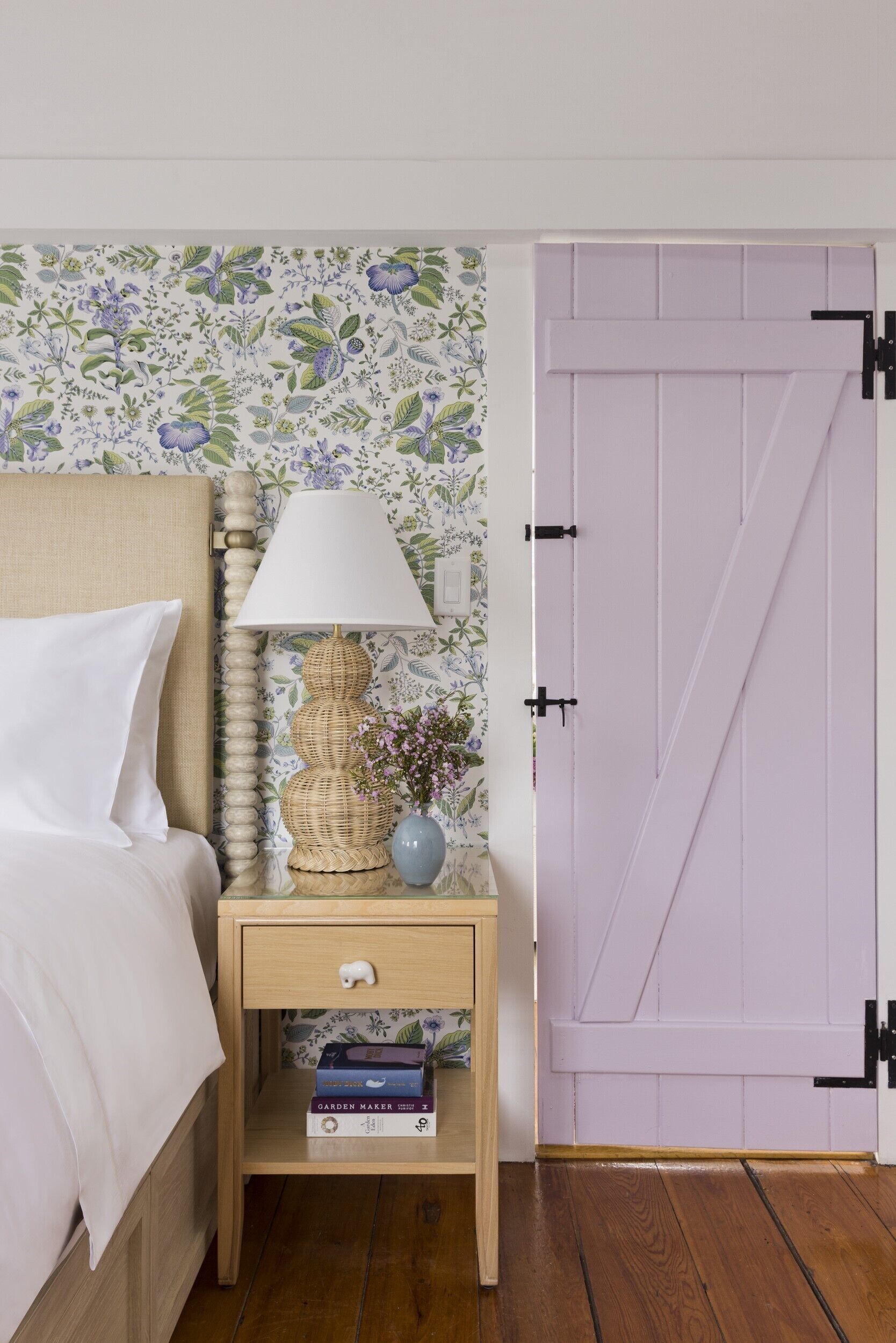 Una cama bien hecha con un almohadón blanco y un cabecero color beige, una mesita de noche con una lámpara, un pequeño florero con flores, y una puerta con un panel morado.