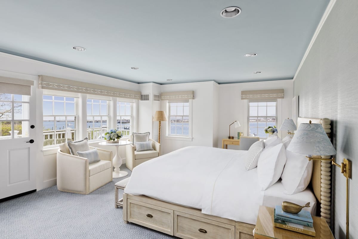 A spacious bedroom with a large bed, a sitting area with a chair and table, and multiple windows offering a view of the sea.
