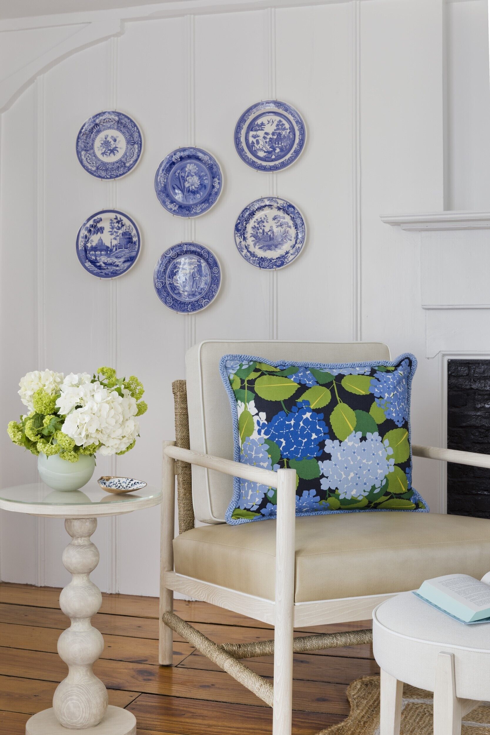 A room with a white chair, a round table with a vase of flowers, and blue and white patterned plates on the wall.