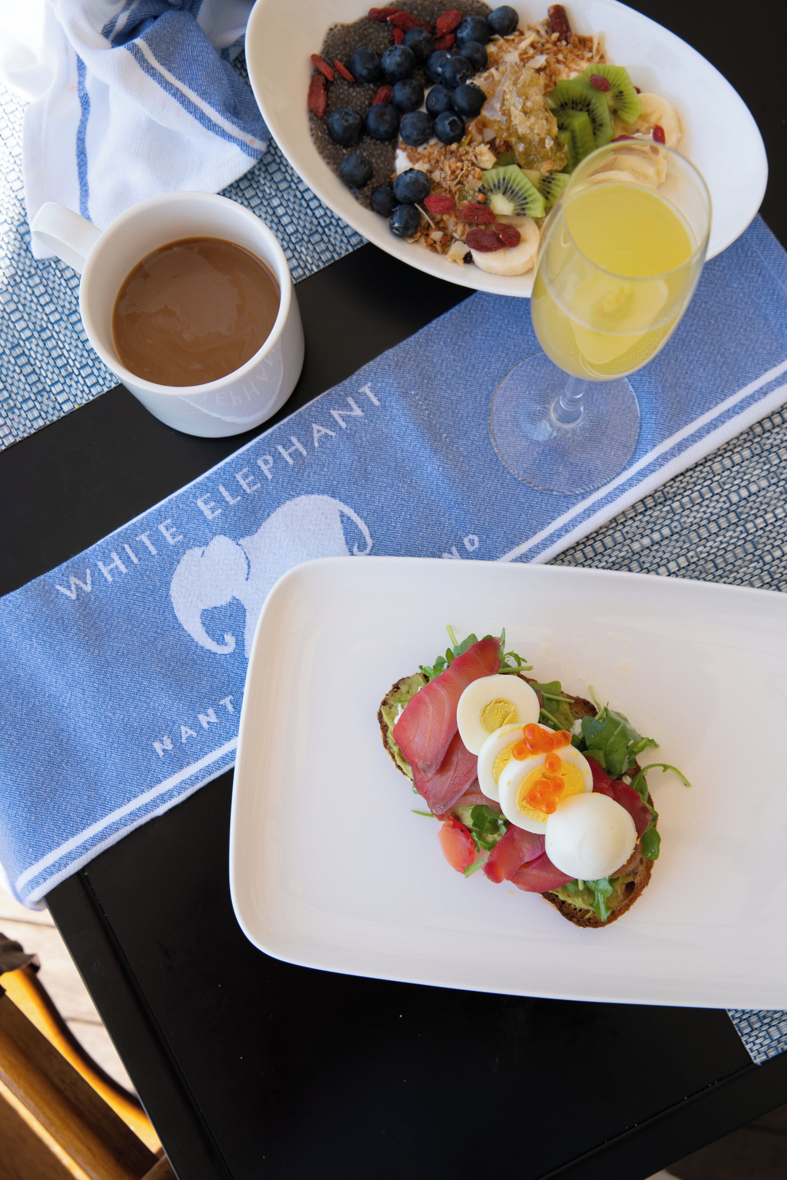 A breakfast setting with a plate of eggs, greens, and salmon, a bowl of granola with fruits, a cup of coffee, and a glass of orange juice on a table with a blue napkin.