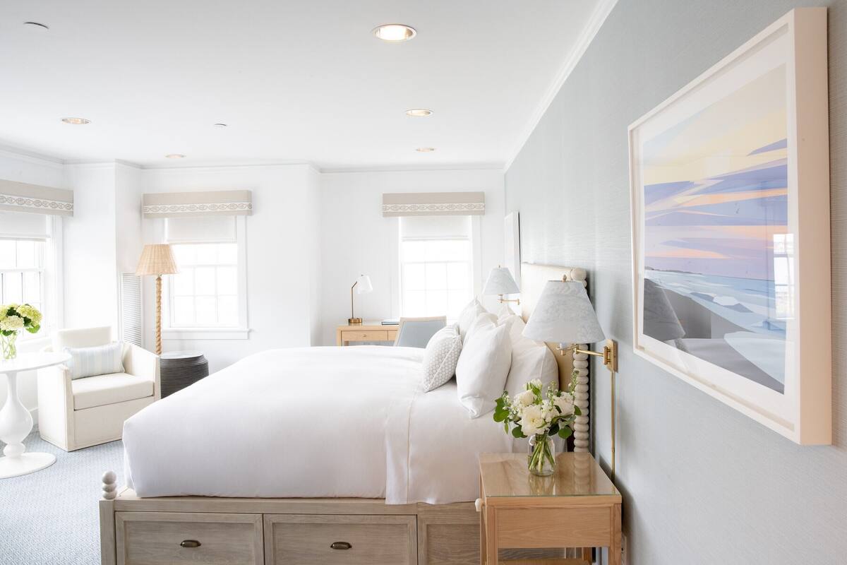 A spacious bedroom with a large bed, bedside lamps, a nightstand, a chair, and a framed picture on the wall.