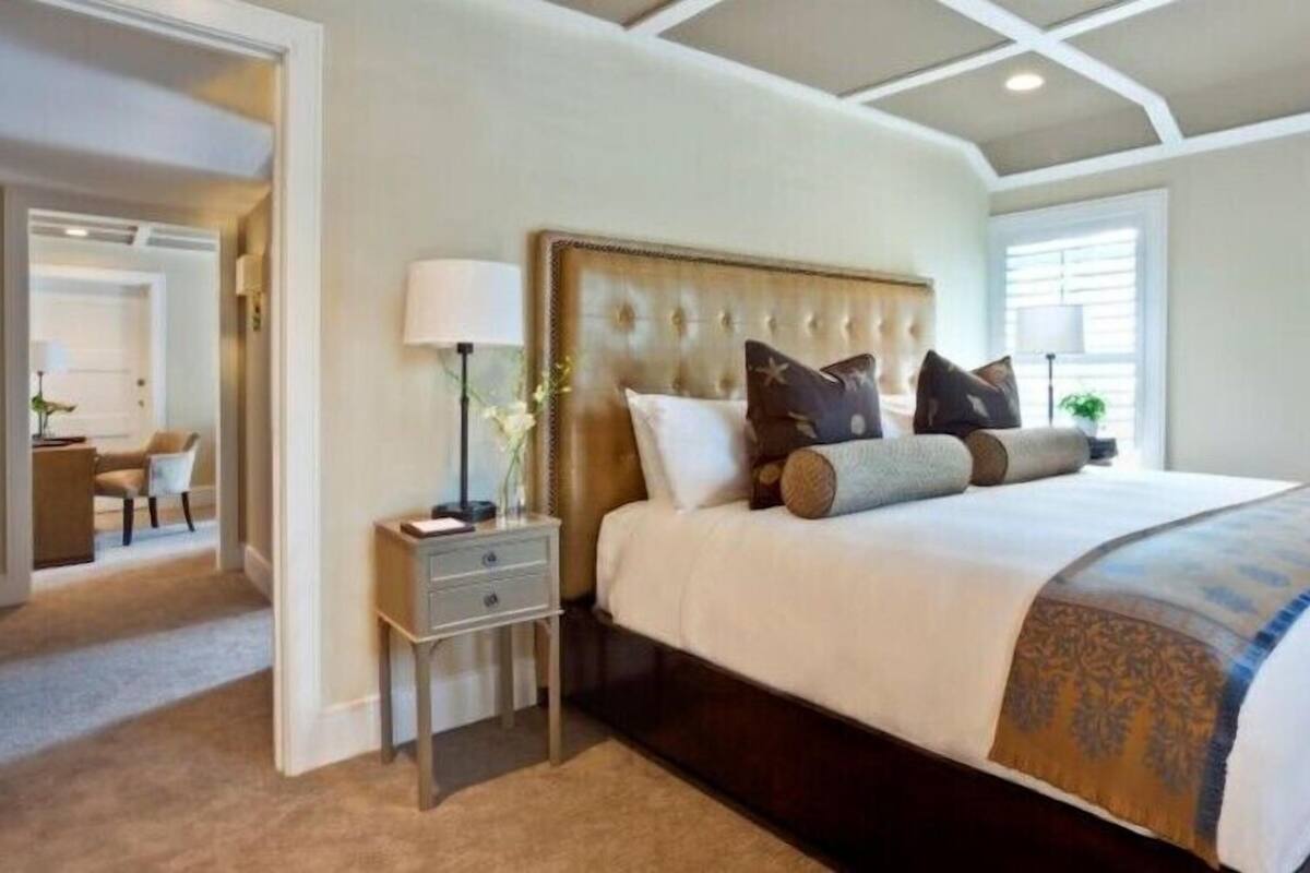 A bedroom with a large bed, a nightstand, a lamp, and a chair in the background.