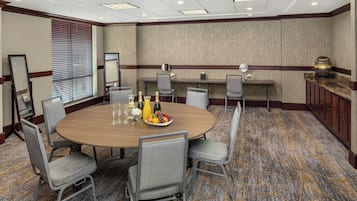 Une salle de conférence avec une table ronde, des chaises grises, un panier de fruits et une bouteille de champagne.
