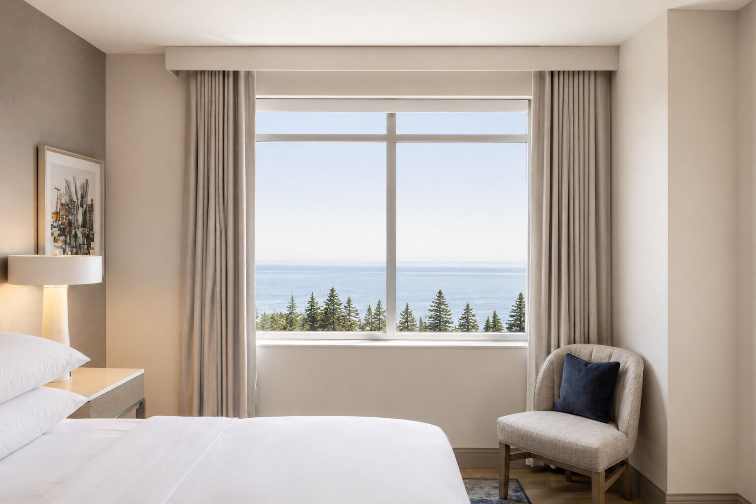 A hotel room with a bed, a chair, a nightstand, a lamp, and a window with a view of the sea and trees.