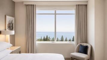 A hotel room with a bed, a chair, a nightstand, a lamp, and a window with a view of the sea and trees.