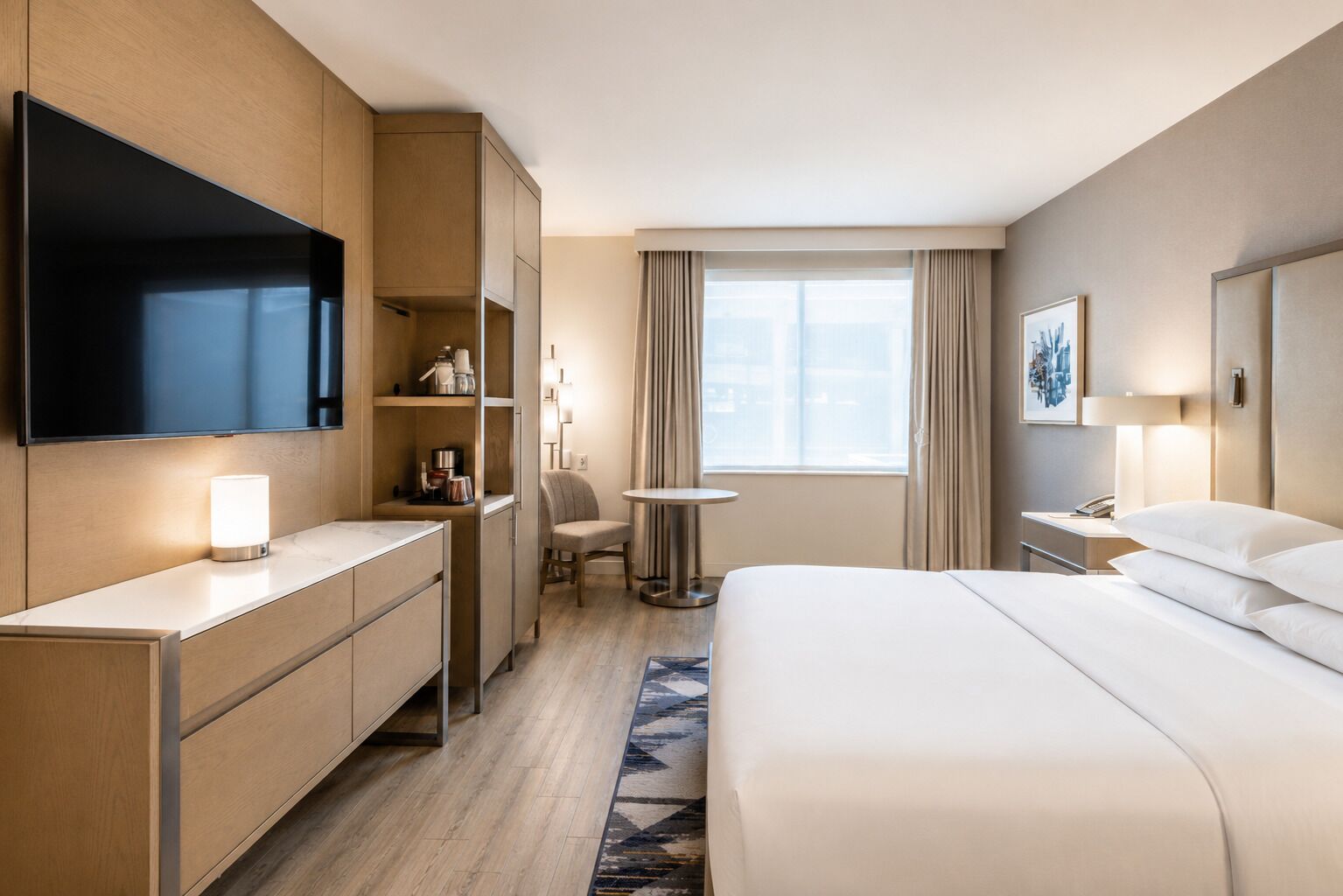 A hotel room with a bed, a desk, a chair, a small table, a TV, and a window with curtains.