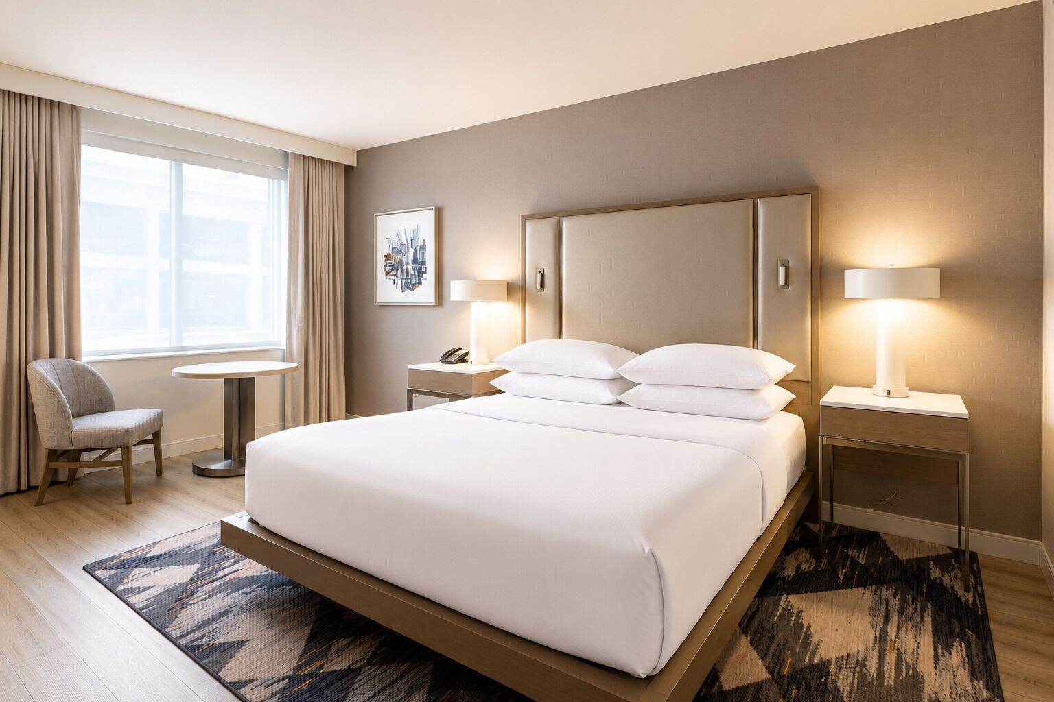 Habitación de hotel con una cama grande, lámparas de noche, una silla, una mesita y una ventana con cortinas.