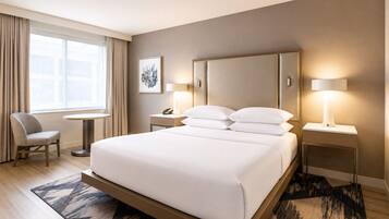 Habitación de hotel con una cama grande, lámparas de noche, una silla, una mesita y una ventana con cortinas.